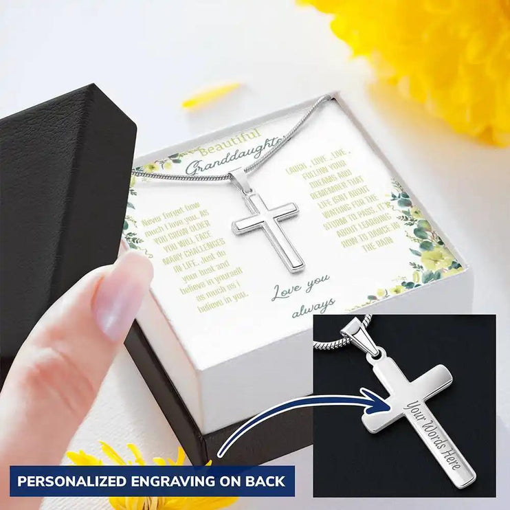 Personalized Cross Necklace with a polished stainless-steel cross and a to granddaughter greeting card in a two-tone box on a table with yellow flowers angle slightly right 