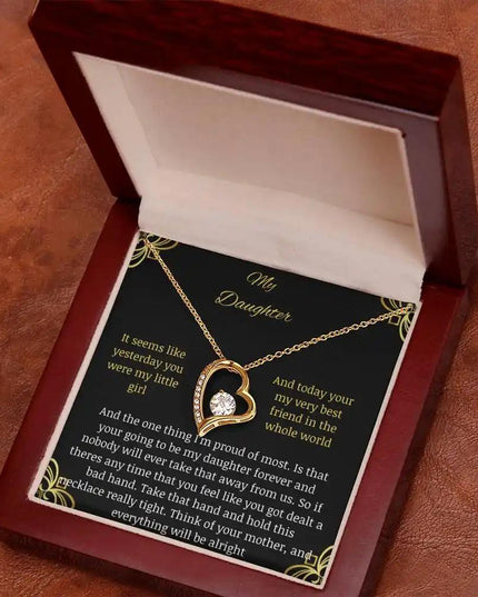 Forever Love Necklace with a yellow gold charm in a mahogany box with a to daughter from mom greeting card on a rustic brown table angled left