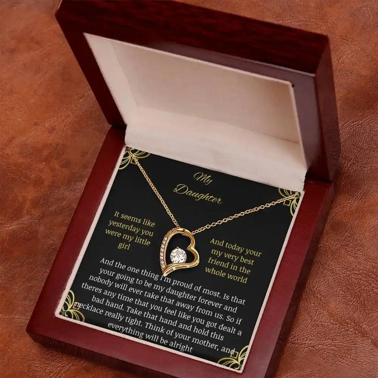 Forever Love Necklace with a yellow gold charm in a mahogany box with a to daughter from mom greeting card on a rustic brown table angled left