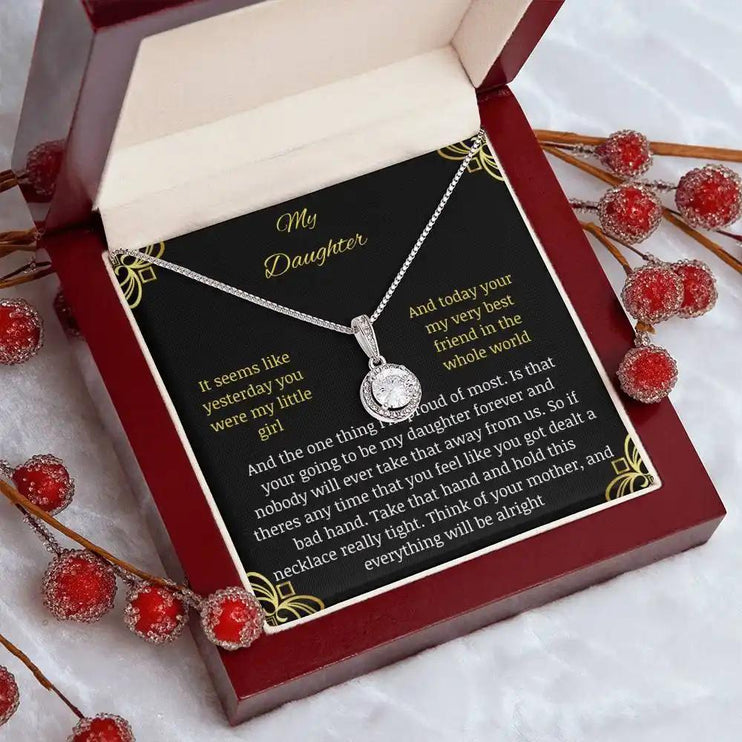 Eternal Hope Necklace in a mahogany box with a to daughter from mom greeting card angled to the right on a white table with red flower beeds