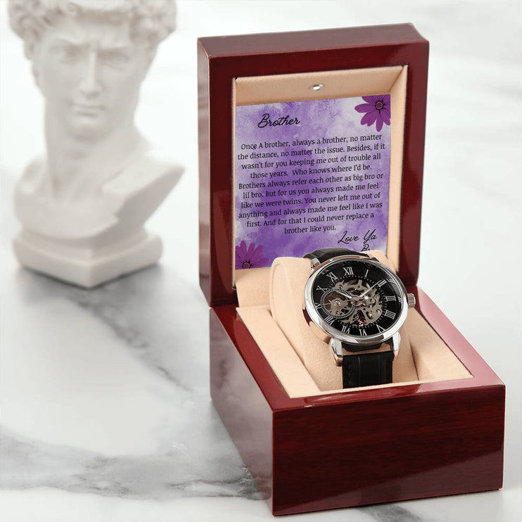 A men's openwork watch and a to brother greeting card in a mahogany box next to a statue.