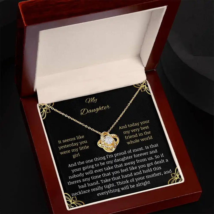 Love Knot Necklace with a yellow gold charm in a mahogany box tilted right with a to daughter from mom greeting card with black background