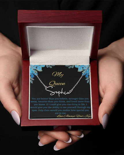 A polished stainless-steel signature name necklace in a mahogany box with a to my queen greeting card being held in hands