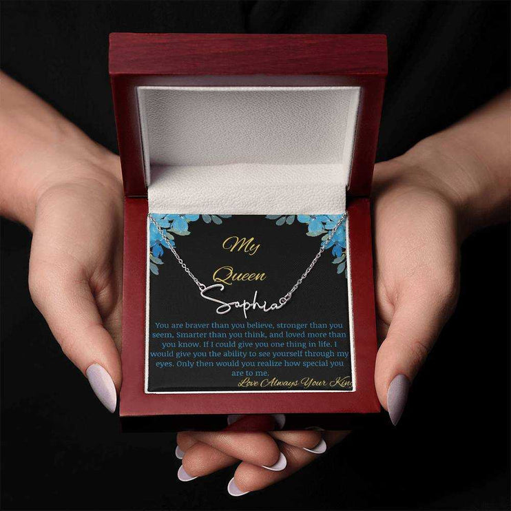 A polished stainless-steel signature name necklace in a mahogany box with a to my queen greeting card being held in hands