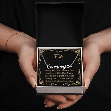 Name Heart Necklace with a polished stainless-steel variant on a To Sis greeting card inside a two-tone box up close view in the hands of a model