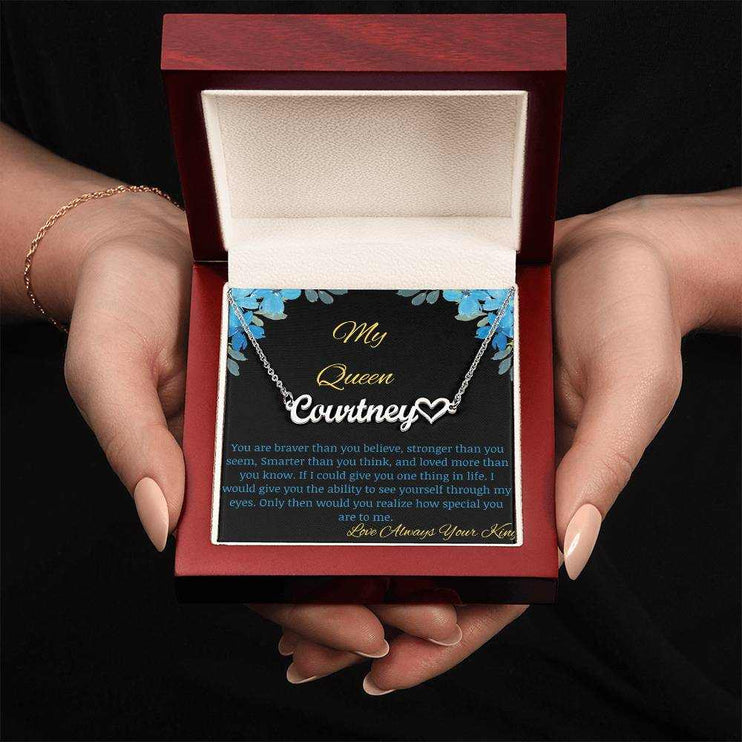 A polished stainless steel name heart necklace in a mahogany box with a to my queen greeting card in a models hands.