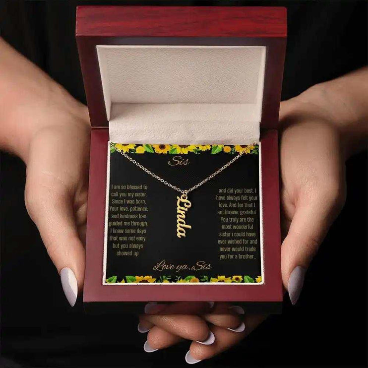 Vertical Name Necklace with a yellow gold finish on a To Sis From Sis greeting card inside a mahogany box close up in models hands