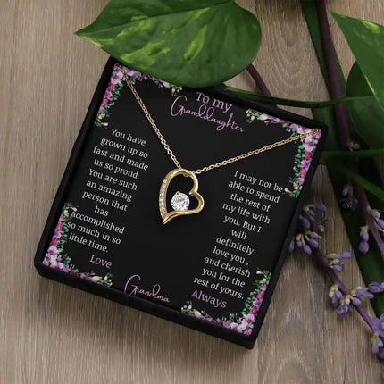 Forever Love Necklace with a yellow gold charm on a for granddaughter from grandpa greeting card in a two-tone box on a table angled left with a green plant beside it