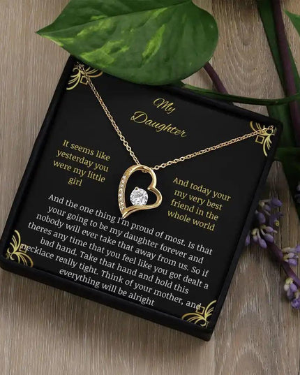 Forever Love Necklace with a yellow gold charm in a two-tone box with a to daughter from mom greeting card sitting on a table angled left with a leaf on corner of box