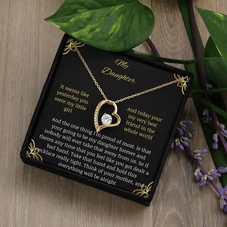 Forever Love Necklace with a yellow gold charm in a two-tone box with a to daughter from mom greeting card sitting on a table angled left with a leaf on corner of box